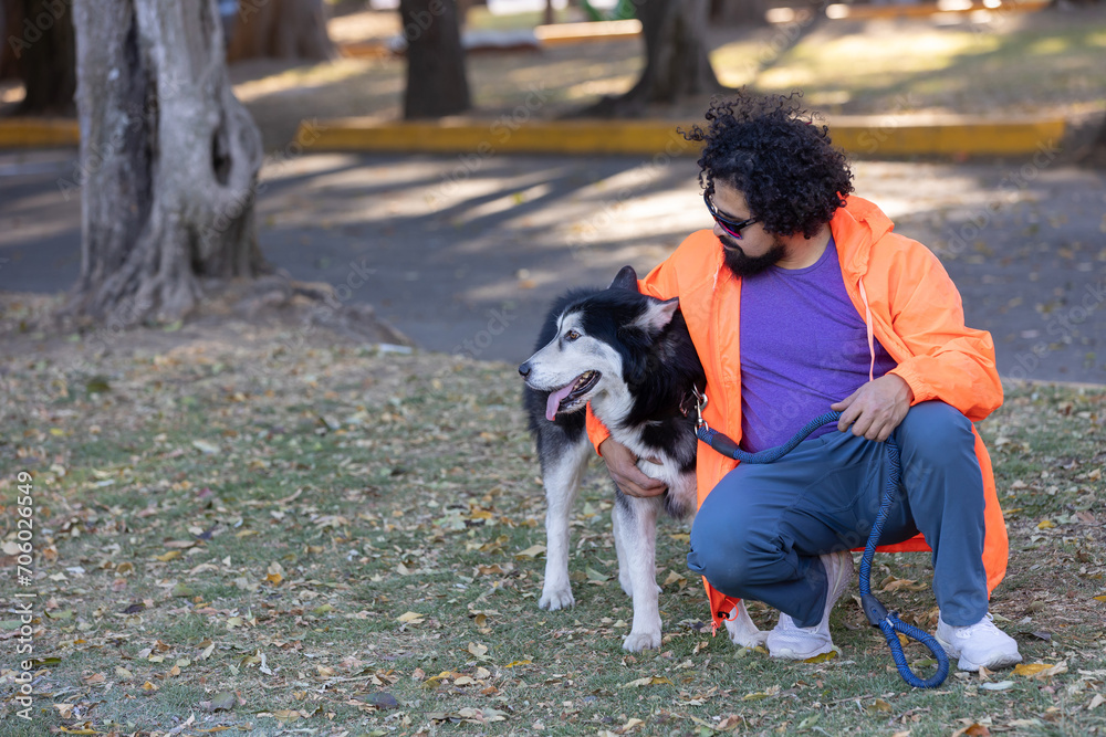 Mexican man with curly hair, beard and wearing casual clothes with sunglasses, sitting in a park hugging his husky dog Mexican man with curly hair, beard and wearing casual clothes with sunglasses, sitting in a park hugging his husky dog