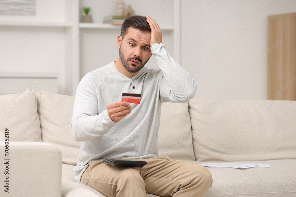 Shocked man with calculator and credit card planning budget at home. Debt problem