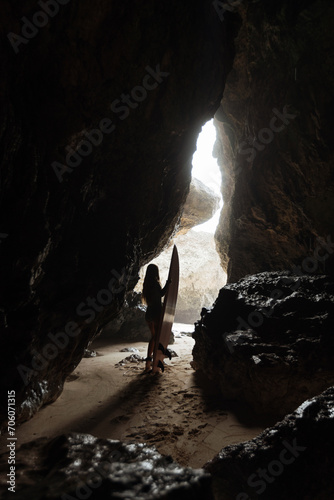 Silhoutte of beautiful young woman surfer in black bikini with surfboard on the beach walk through the cave to the seashore on Bali island   Uluwatu