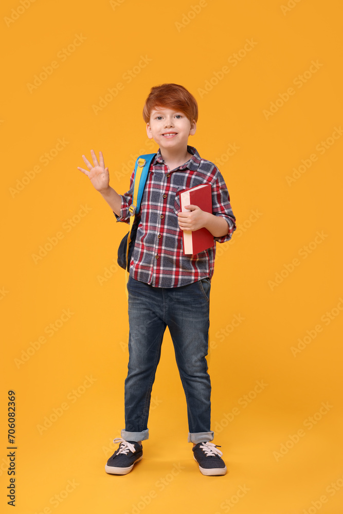 Happy schoolboy with backpack and book waving hello on orange background Happy schoolboy with backpack and book waving hello on orange background