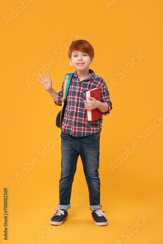 Happy schoolboy with backpack and book waving hello on orange background