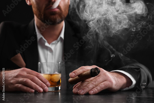 Man with glass of whiskey smoking cigar at dark marble table on black background, closeup