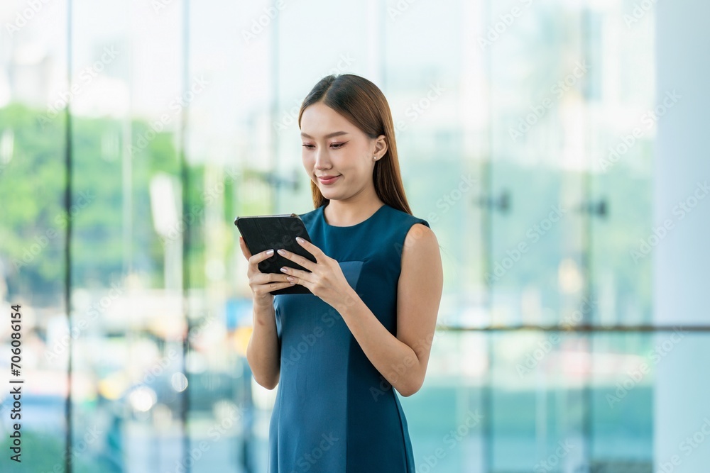 Fototapeta premium Young Asian Woman Smiles Happily While Using Digital Tablet to Manage Her Tasks Remotely While Out Of Office