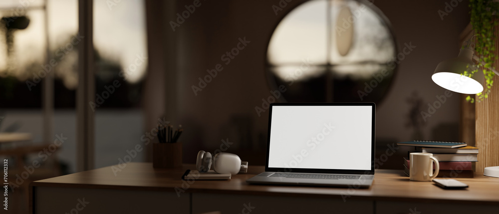 Naklejka premium A cosy, modern home workspace at night with a white-screen laptop computer mockup on a wooden desk.