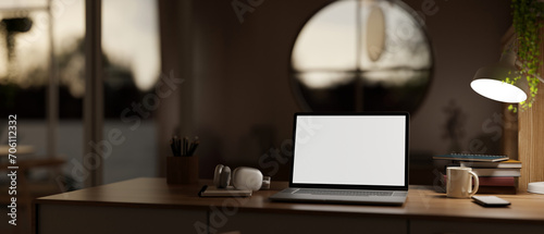 A cosy, modern home workspace at night with a white-screen laptop computer mockup on a wooden desk.