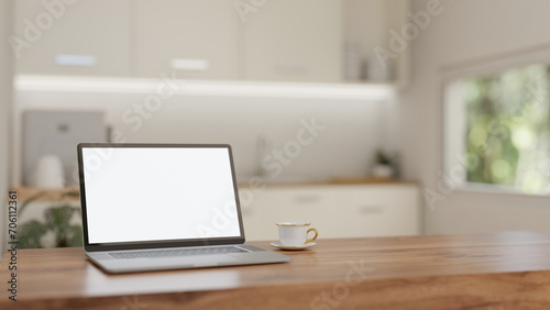 A white-screen laptop mockup and a coffee cup on a wooden table in a minimalist white kitchen.