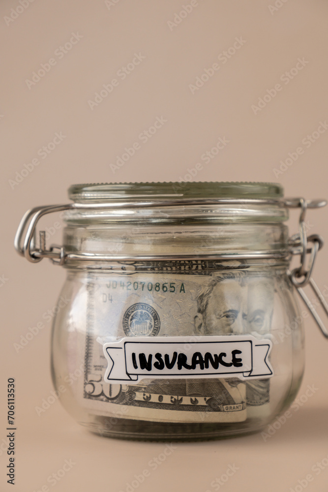 Saving Money In Glass Jar filled with Dollars banknotes. INSURANCE transcription in front of jar. Managing personal finances extra income for future insecurity. Beige background Saving Money In Glass Jar filled with Dollars banknotes. INSURANCE transcription in front of jar. Managing personal finances extra income for future insecurity. Beige background