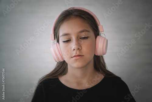 Serious displeased Caucasian kid girl wearing black dress on grey background being angry wears stereo headphones listens music photo