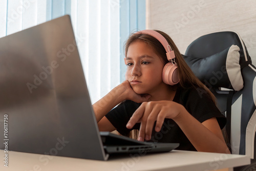 Portrait of little girl in wireless headset using laptop, studying online at home, interested happy student typing on keyboard looking at pc screen, watching webinar, online course, doing homework