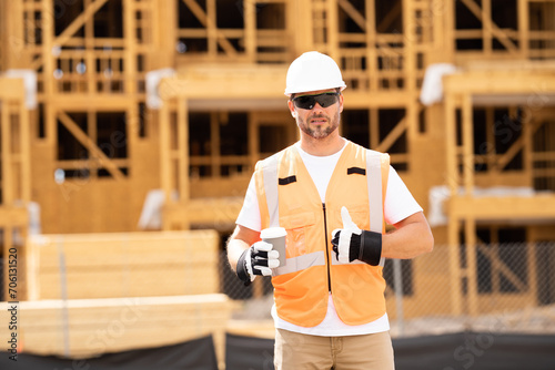 Coffee, engineer and construction worker relax on break at construction site. handsome male builder in hard hat smiling at camera. Construction Worker on Duty. Contractor and the Wooden House Frame