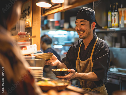 A candid shot of a japanese chef, who's also the shop owner, offering ramen as he hands a customer their order in his ramen restaurant