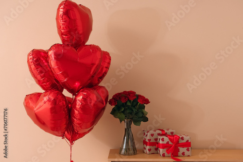 Beautiful bouquet of roses with heart-shaped balloons and gift boxes on commode near beige wall. Valentine's Day celebration