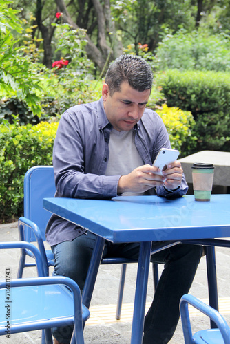 40-year-old dark-skinned Latino man drinks a coffee and uses his cell phone on a park bench to work remotely, make calls, send messages, buy and pay online