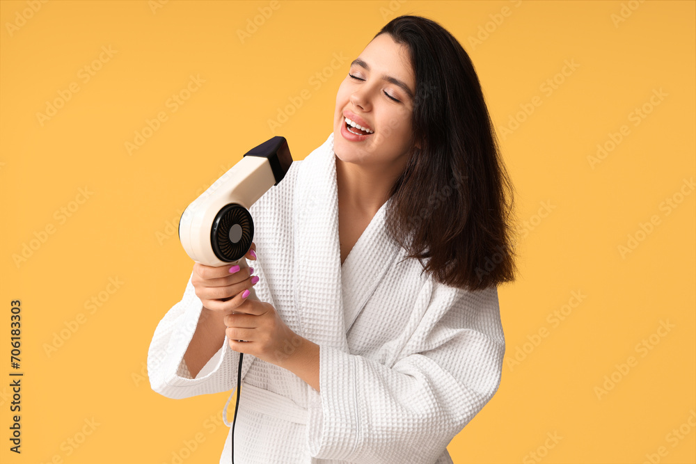 Beautiful young happy woman in bathrobe with hair dryer on yellow background Beautiful young happy woman in bathrobe with hair dryer on yellow background