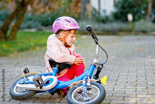 Cute little girl sitting on the ground after falling off her bike. Upset crying preschool child with safe helmet getting hurt while riding a bicycle. Active family leisure with kids.