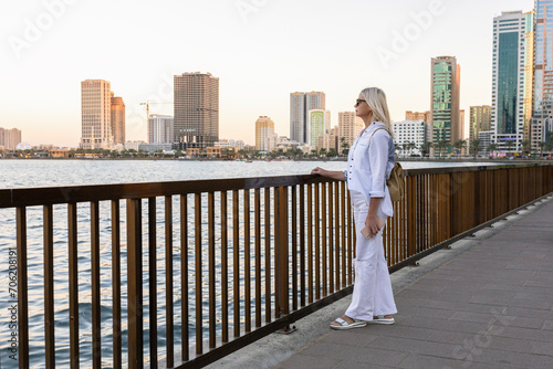 Stylish woman standing on the waterfront promenade and enjoying beautiful cityscape at sunset.