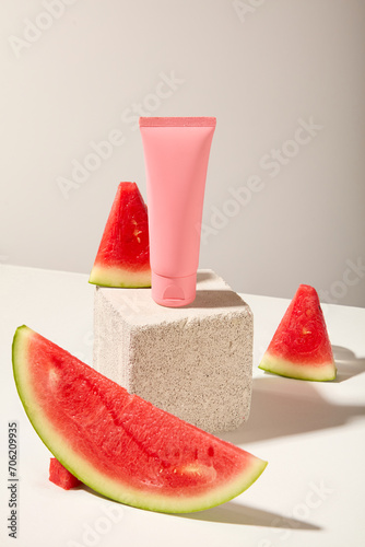 On a white background, a pink plastic tube displayed on stone podium with fresh juicy watermelon slices. Front view, mockup scene for advertising cosmetic of watermelon ingredient