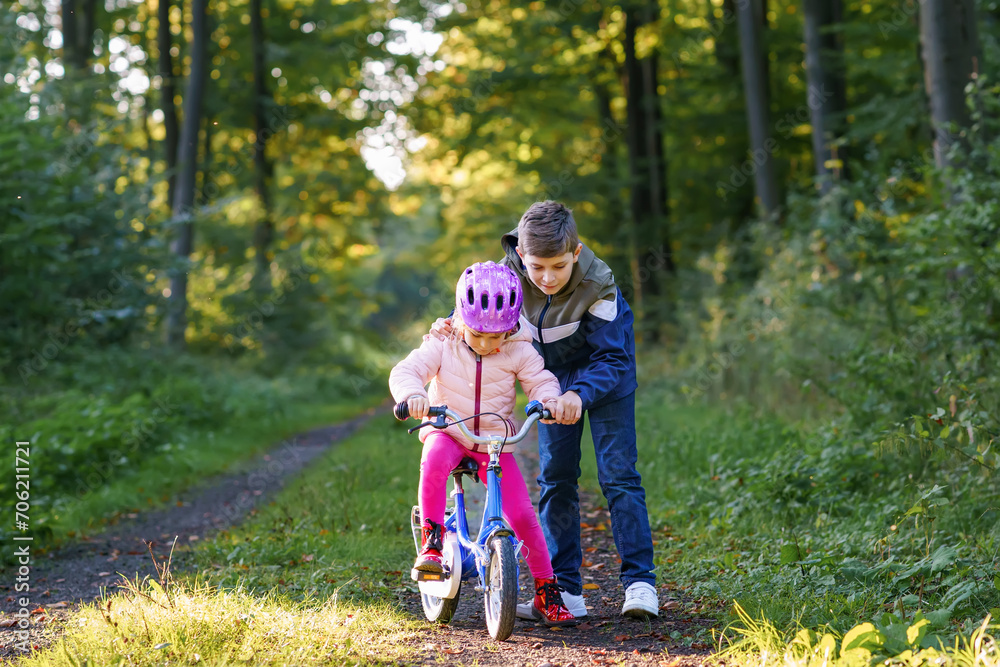 Cute little preschool girl in safety helmet riding bicycle. School kid boy, brother teaching happy healthy sister child cycling and having fun with learning bike. Active siblings family outdoors. Cute little preschool girl in safety helmet riding bicycle. School kid boy, brother teaching happy healthy sister child cycling and having fun with learning bike. Active siblings family outdoors.