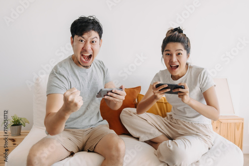 Happy asian Thai couple enjoy playing game together, glad celebrating victory with raising hands, woman showing mobile phone to man, sitting on sofa spending time together in room apartment.