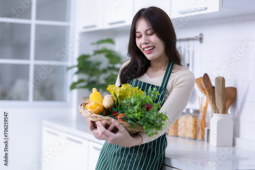Obraz na płótnie Happy portrait of young asian woman holding a basket of vegetables of standing a cheerful preparing food and enjoy cook cooking with vegetables, while standing on a kitchen Condo life or home w salonie