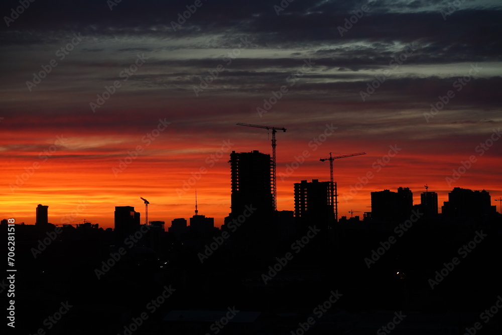 Sunset with orange and red sky in cityscape with silhouettes of buildings. Sunset against the backdrop of a large construction site with several working cranes and unfinished high-rise buildings