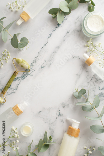 Natural body care ensemble. Top view vertical shot featuring gua sha roller, skincare essentials, cream and serum bottles, eucalyptus, and gypsophila bloom on a marble background with space for text