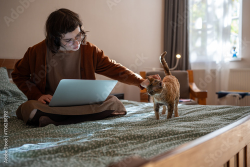 Busy teenager pet owner sitting on bed, working on laptop computer as freelancer. Beloved purring ginger breed cat Devon Rex diverting girl attention, requiring to stroke, caress, amuse, play