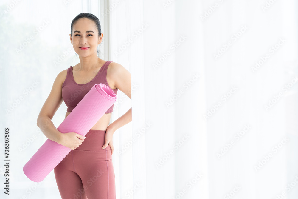 Happy young beautiful asian fitness woman standing pose holding yoga mat. Cheerfully sporty female workout and exercise wearing sportswear. Charming woman relax after fitness training.