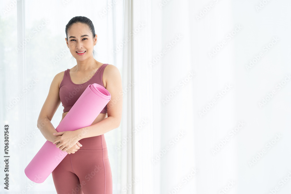 Obraz premium Happy young beautiful asian fitness woman standing pose holding yoga mat. Cheerfully sporty female workout and exercise wearing sportswear. Charming woman relax after fitness training.