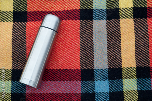 Winter Adventure: Classic White Stainless Steel Thermos Rests on a Checkered Colorful Rug, Providing Warmth and Comfort in the Chilly Outdoors