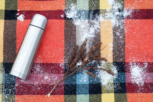 Top view of a classic white stainless steel thermos on a checkered colorful checkered rug dusted with snow and a dry plant.