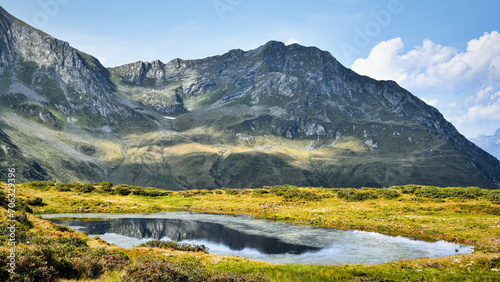 Bergsee mit Reflexion, grüne Wiese, Berge und Felsen im Hintergrund, blauer Himmel mit Wolken