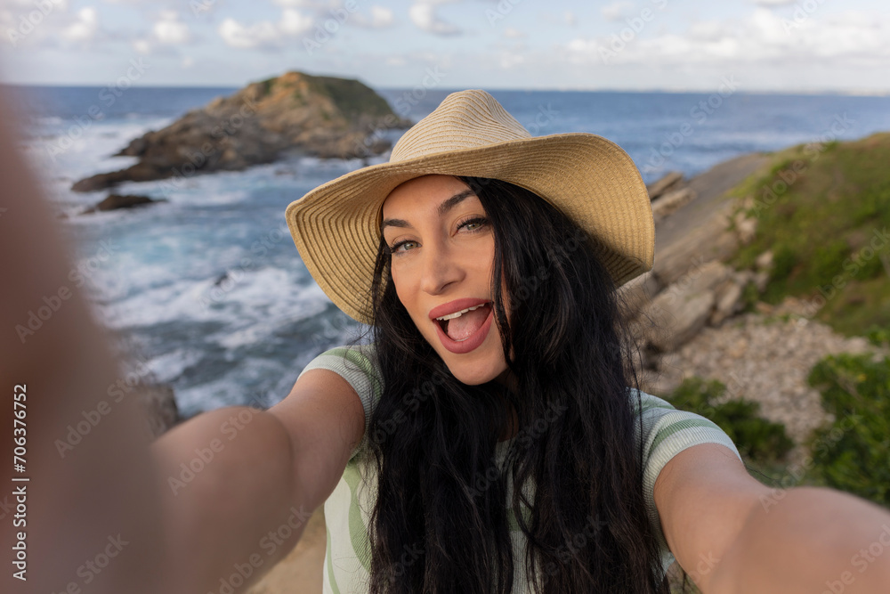 beautiful woman with hat very smiling taking a selfie with her smartphone on her vacation trip with the sea in the background