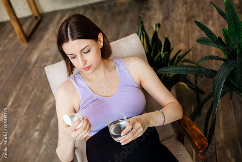 Young happy pregnant woman taking prenatal vitamins, holding glass of water and pill while sitting at armchair at home