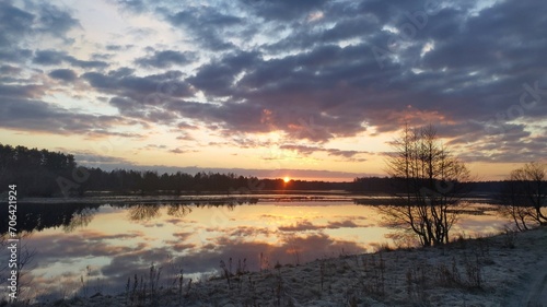 The sun rises from behind the forest on the far side of the lake and brightly colors the cloudy sky. Trees and clouds are reflected in the water. The grass on the shore is covered with frost