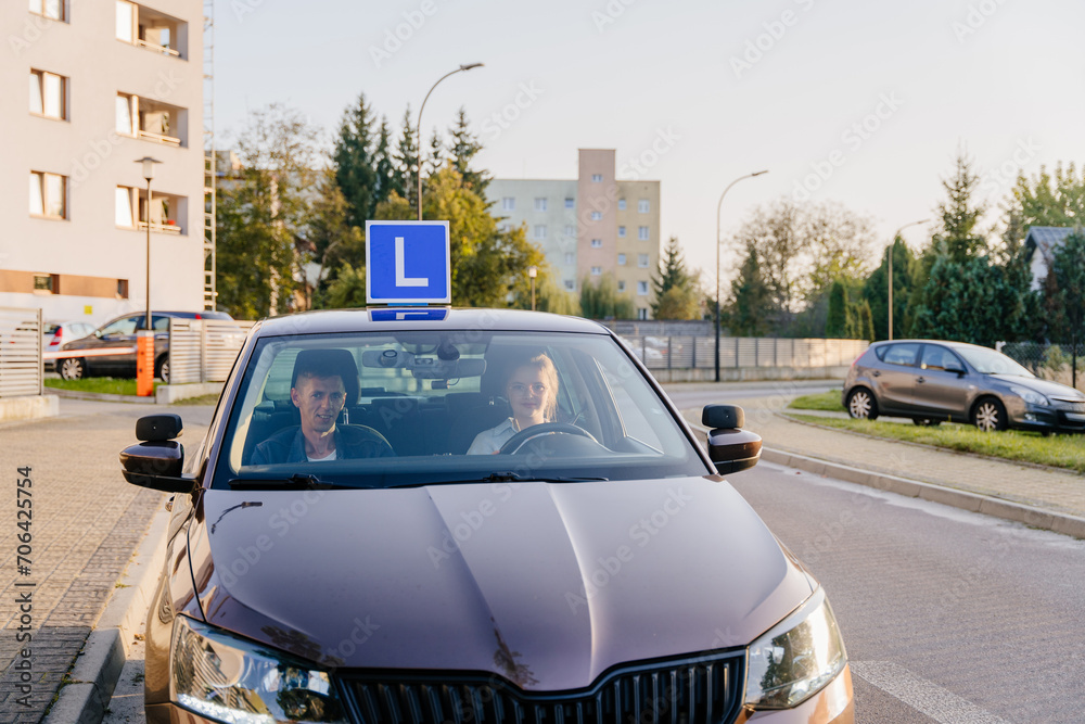 Brown car with blue L plate on a roof driving down a city street with an instructor and a young female student inside. Young Teen Girl Doing Driving Exam with Examiner.