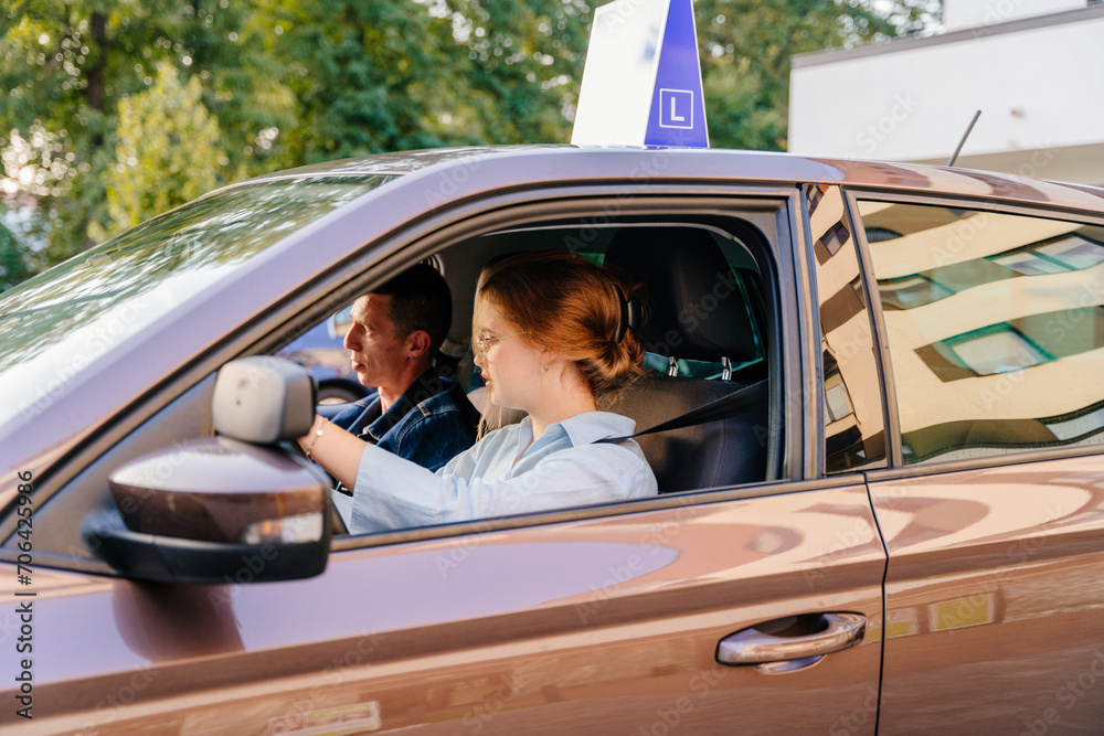 Carefully instructor sitting in a learning car next to a female student driver. Happy woman drives brown left-hand driving vehicle with blue L plate on a roof.