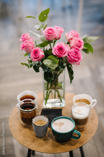 A coffee table with various drinks. Cups with tea, coffee, cappuccino, and latte stand on round wooden table. Pink bouquet of rose flowers in vase on blurry background in a white living room interior.