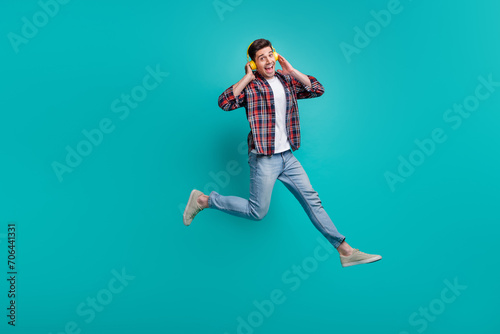 Full length photo of cool funky man dressed plaid shirt jumping high enjoying music headphones isolated turquoise color background
