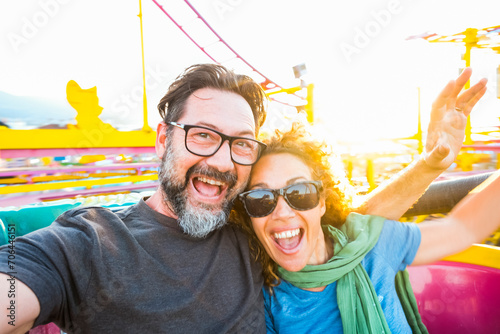 Adult couple have fun together on a roller coaster in amusement park taking selfie picture with the phone. People enjoying holiday lifestyle. Mature man and woman enjoy time outdoor leisure activity