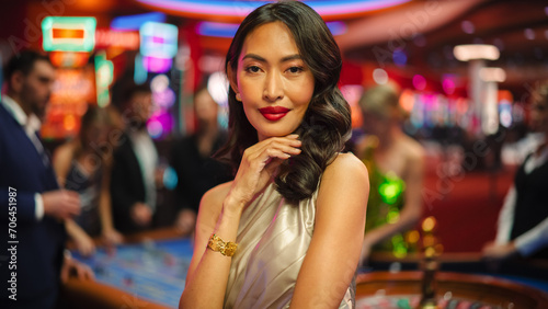 Portrait of a Sophisticated Asian Woman in a Stunning Dress Posing, Smiling, Looking at Camera in a Glamorous Casino with Gamblers Playing Roulette in Colorful Bokeh Background