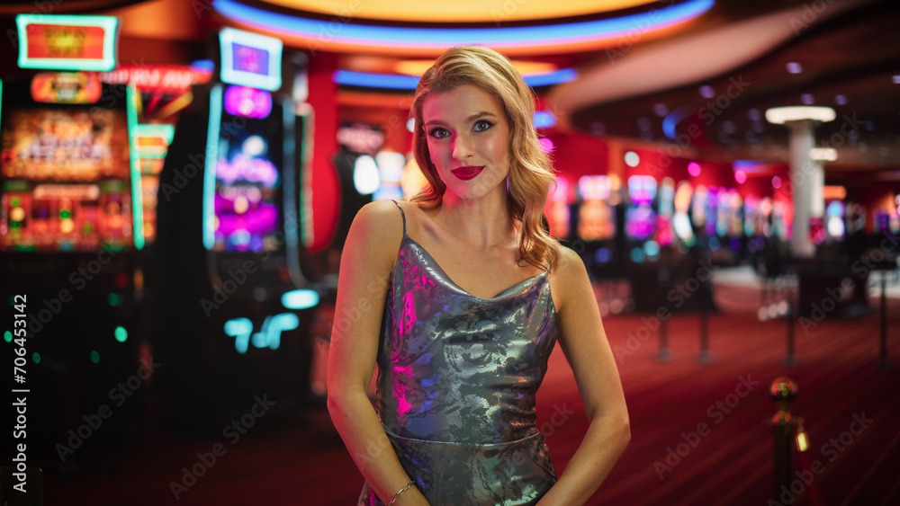 Fototapeta premium Beautiful Young Caucasian Woman Posing In Prestige Casino Next To Baccarat Table. Charismatic Female Croupier Smiling, Looking At Camera And Wearing Fancy Silver Dress