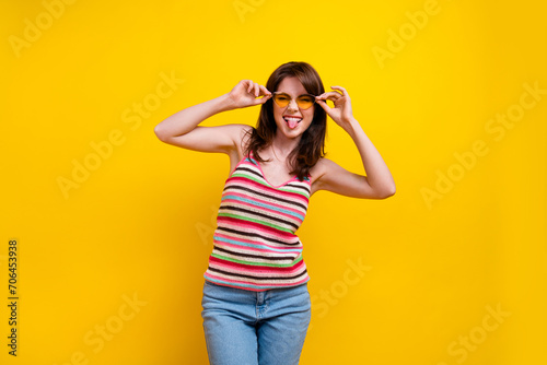 Photo of lovely positive eccentric woman dressed knitwear top touching sunglass showing tongue isolated on vibrant yellow color background