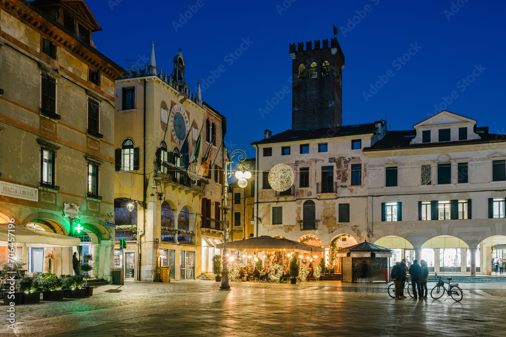 Bassano del Grappa, Italy - January 09, 2024: People Piazza Libertà in cafe tables in the evening in winter. On the left the town hall.