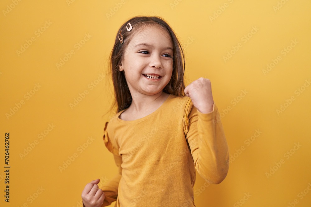 Adorable hispanic girl joyfully standing, arms raised in victorious celebration of success on a yellow isolated background, eyes softly closed, yes! Adorable hispanic girl joyfully standing, arms raised in victorious celebration of success on a yellow isolated background, eyes softly closed, yes!