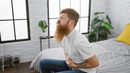 Tormented young adult redhead guy touching his aching stomach while sitting miserably on bed in bedroom, suffering from stomach pain, an indoor awakening ordeal photo