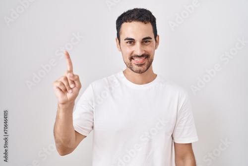 Handsome hispanic man standing over white background showing and pointing up with finger number one while smiling confident and happy.