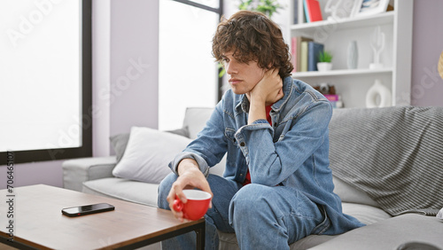 Thoughtful young man with curly hair, holding a red mug in a modern living room while sitting on a gray couch near a smartphone.