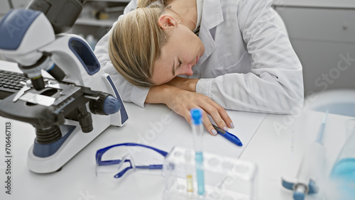 A tired woman rests her head on a lab table next to a microscope and test tubes, depicting exhaustion in a scientific setting.