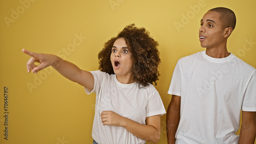 Stunned and gorgeous, this cool couple is pointing to the side together in surprise on a fun-loving yellow isolated background